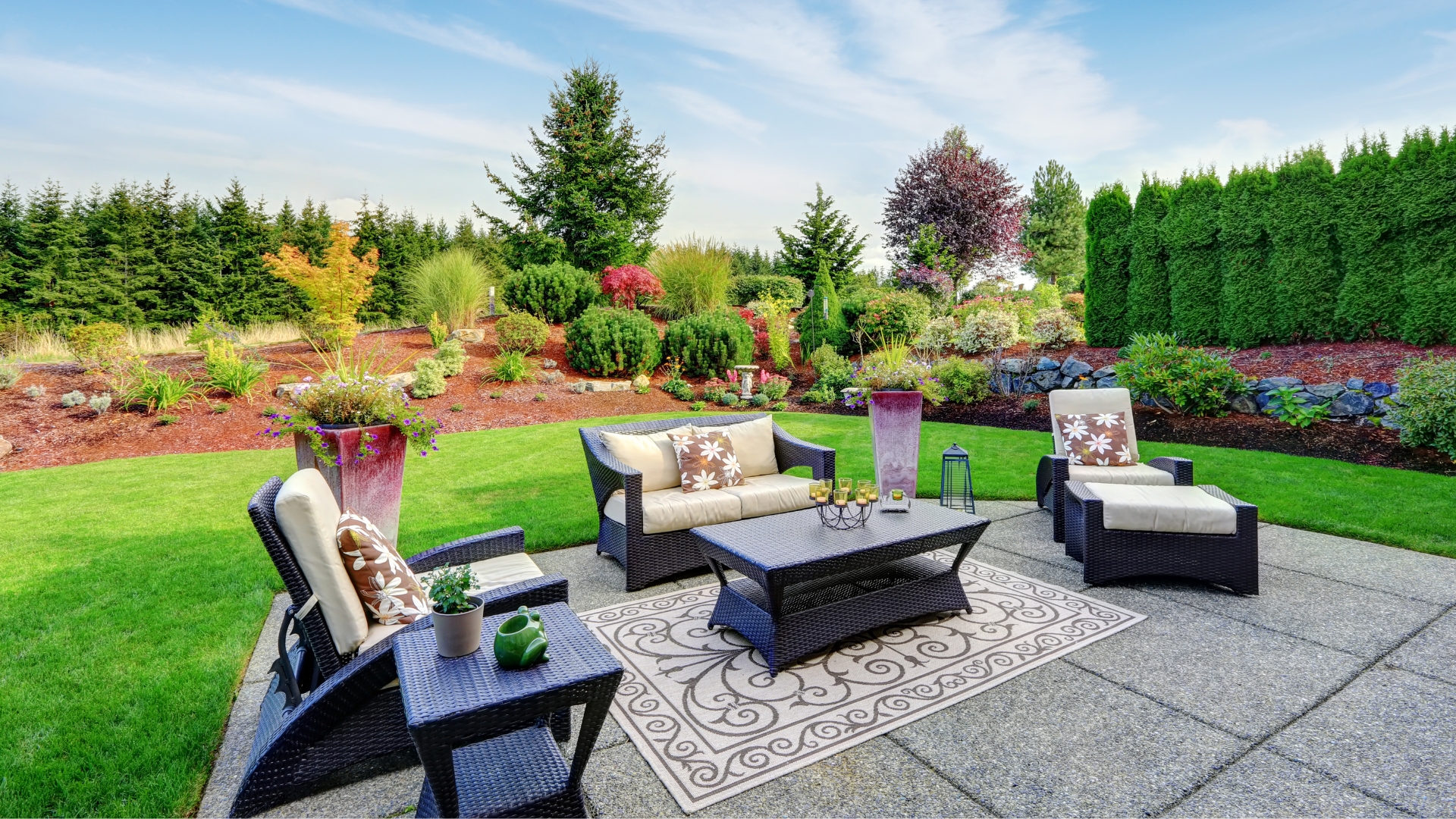 Beautifully landscaped backyard with vibrant shrubs and evergreens, featuring a concrete patio with wicker furniture, outdoor rug, and decorative planters.