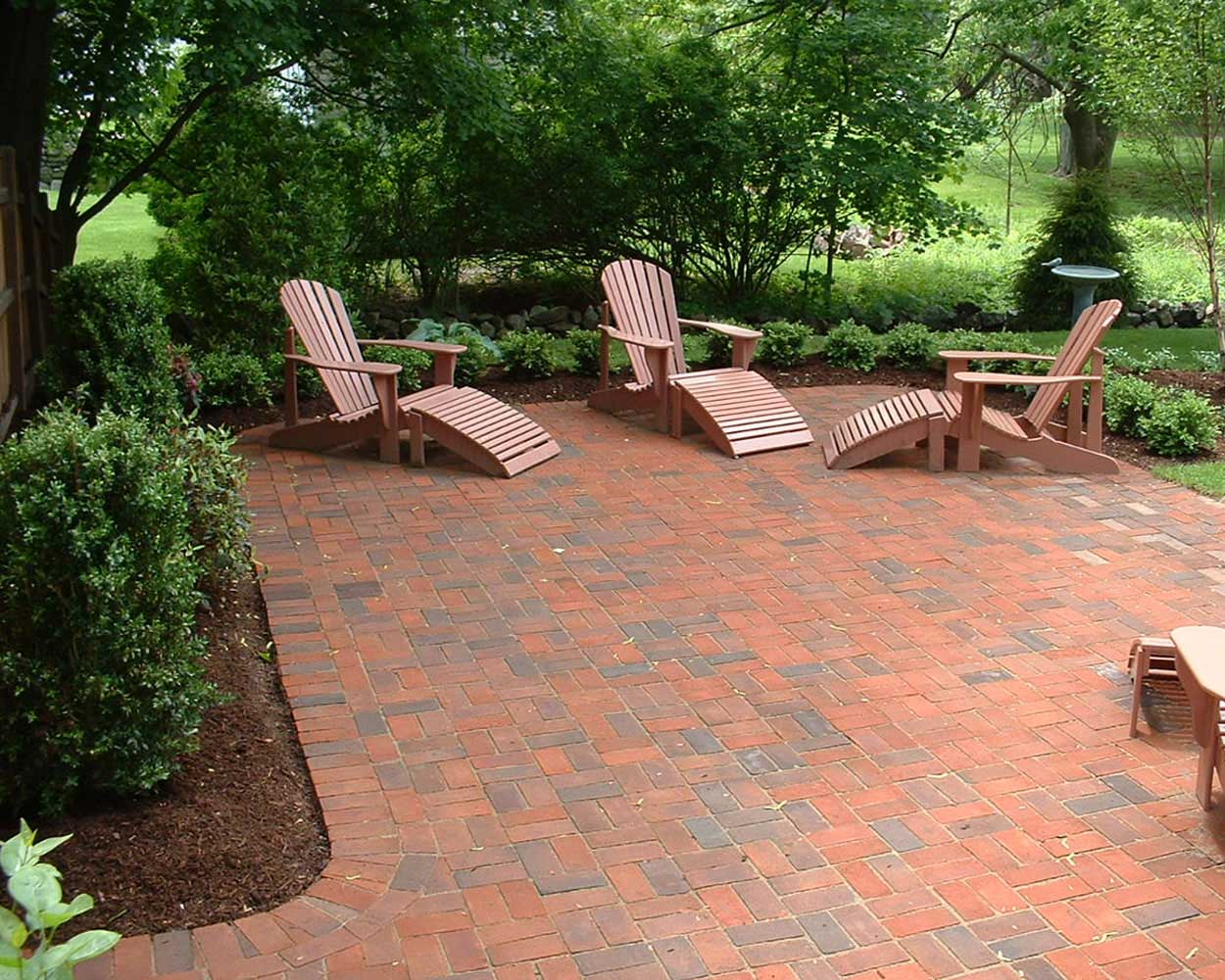 Brick patio with wooden Adirondack chairs and curved footrests, surrounded by neatly edged garden beds and lush green landscaping in a shaded backyard setting.