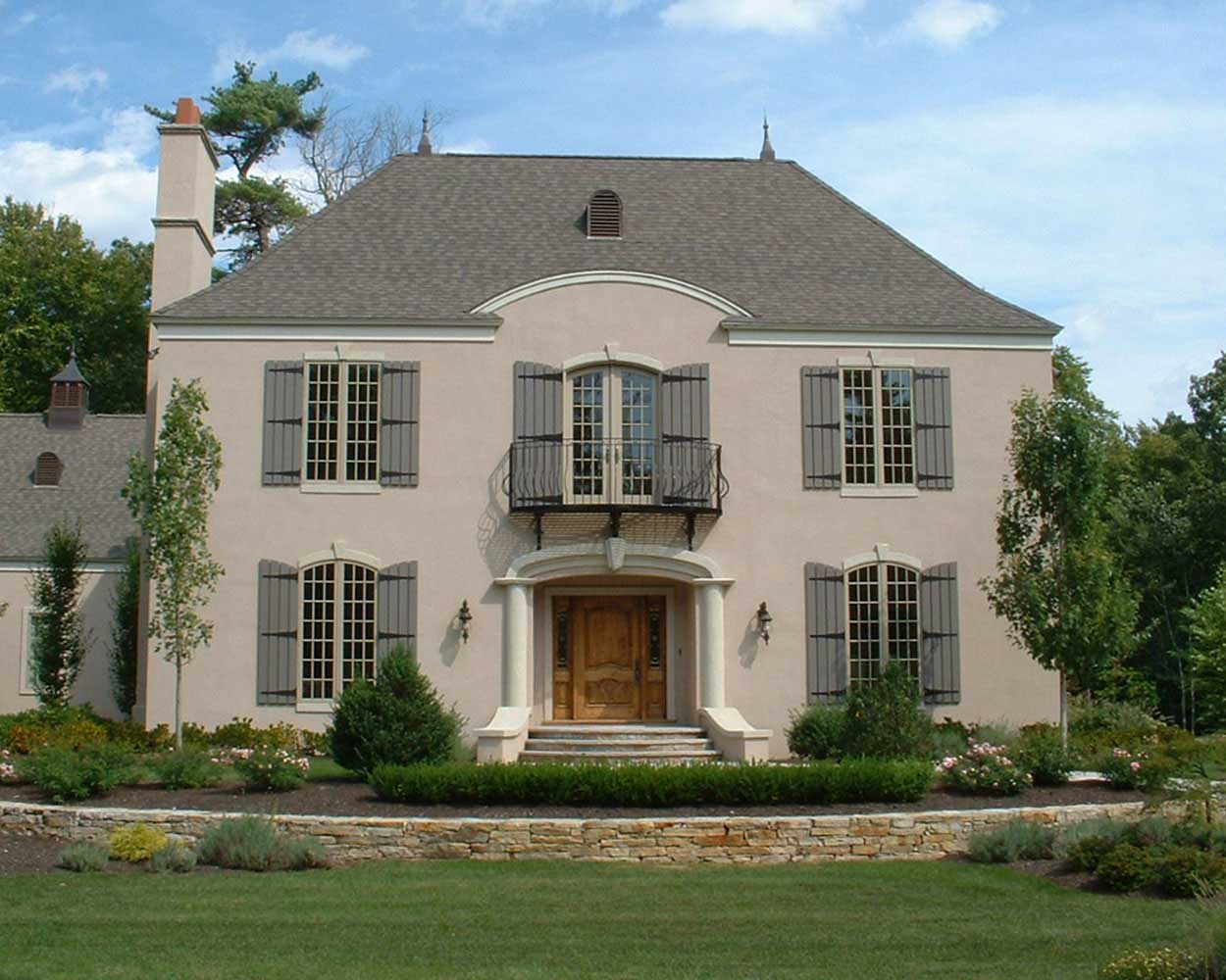 Elegant stucco French-style home with symmetrical windows, wrought-iron balcony, wooden double doors, and a terraced front yard with stone retaining wall and formal landscaping.